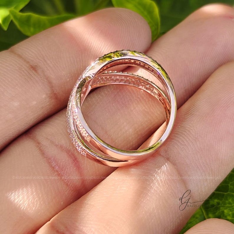May include: A rose gold-toned ring featuring a multi-band crossover design. The surface of the bands is encrusted with numerous small, clear, sparkling stones that catch the light. The ring has a polished metallic finish and a wide, layered structure.