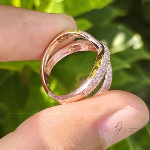 May include: A rose gold crossover ring featuring two intersecting bands, with one band fully encrusted in small, clear pav&eacute;-set stones. The metal has a polished finish, and the design creates an open, layered appearance on the finger.