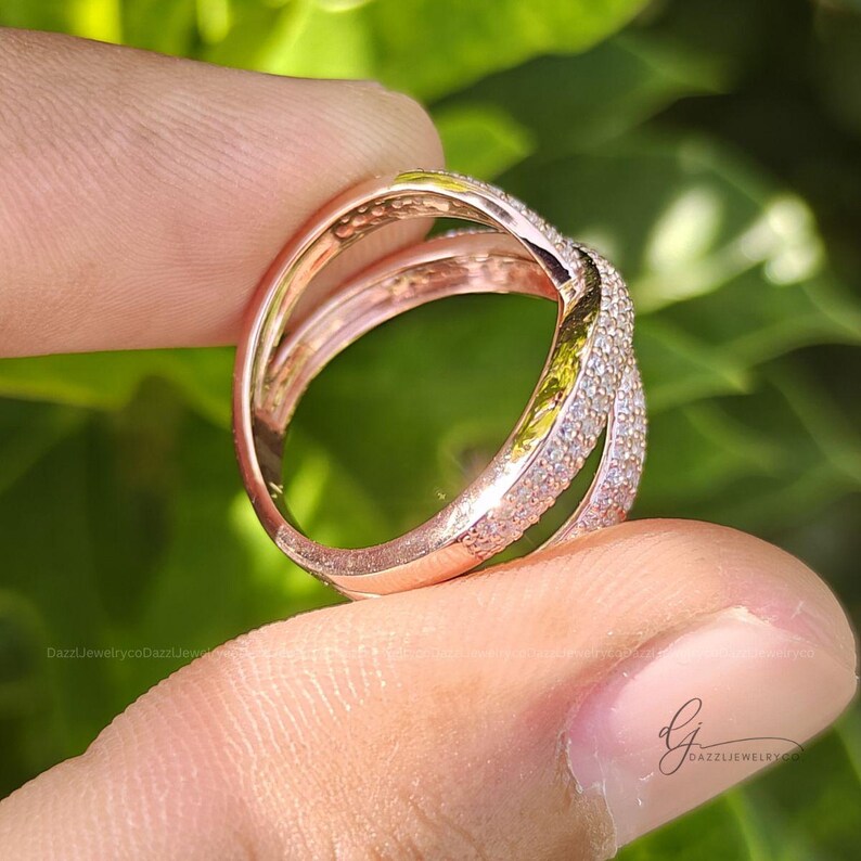 May include: A rose gold crossover ring featuring two intersecting bands, with one band fully encrusted in small, clear pav&eacute;-set stones. The metal has a polished finish, and the design creates an open, layered appearance on the finger.