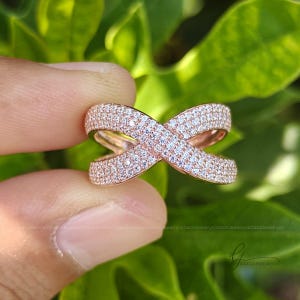 May include: A rose gold crossover ring featuring a crisscross design completely encrusted with small, clear, sparkling pav&eacute;-set stones. The band has a wide, open structure that forms an X shape, showcasing a metallic finish and numerous reflective white stones.