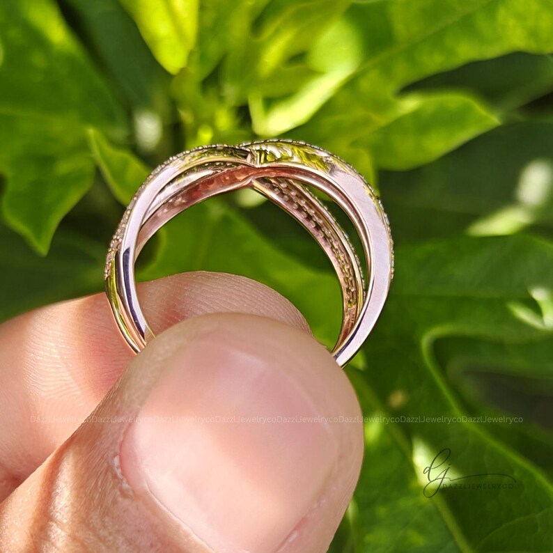 May include: A rose gold crossover ring featuring two intertwined bands. One band is smooth and polished, while the other is encrusted with small, clear, sparkling stones along its entire visible surface, creating a layered and textured appearance.