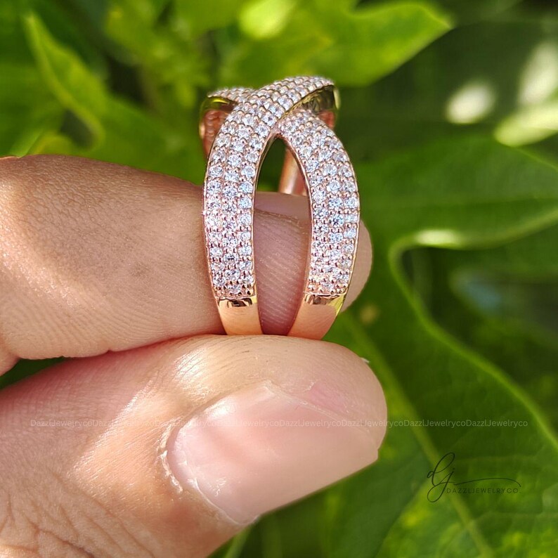 May include: A rose gold crossover ring featuring two intersecting bands. The surface of the bands is densely encrusted with numerous small, clear, sparkling stones that cover the entire visible exterior of the metal structure.
