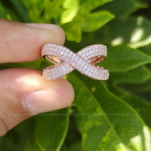 May include: A rose gold ring featuring an X-shaped crossover design. The entire surface of the band is encrusted with numerous small, clear, sparkling stones that cover the metal, creating a continuous shimmering effect across the crisscross structure.
