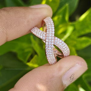 May include: A rose gold ring featuring an infinity-shaped design, fully encrusted with numerous small, clear, sparkling stones. The band curves into a continuous loop, creating a symmetrical crossover pattern that catches the light.