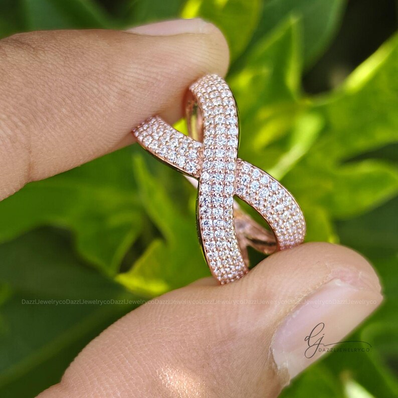 May include: A rose gold ring featuring an infinity-shaped design, fully encrusted with numerous small, clear, sparkling stones. The band curves into a continuous loop, creating a symmetrical crossover pattern that catches the light.