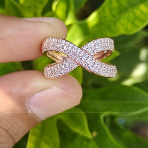 May include: A rose gold crossover ring featuring an X-shaped design. The entire surface of the band is encrusted with numerous small, clear, sparkling stones that cover the metal, creating a continuous shimmering effect across the entire piece.