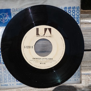Può includere: Un disco in vinile nero con il testo "ENTREGA LA PALABRA" e "WAR" stampato sull'etichetta. Il disco è nella sua custodia originale, che presenta la parola "DISCOS" ripetuta in blu.