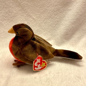 May include: A soft toy robin bird with brown and tan plumage, a red breast, and a yellow beak. The bird has a tag attached to its left side. The toy is set against a white, textured background.