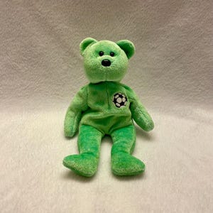 Puede incluir: Un oso de peluche verde con una nariz negra y un emblema de balón de fútbol en el pecho. El oso está hecho de un material suave y difuso y tiene la forma clásica de un oso de peluche. El oso está sobre una superficie blanca.