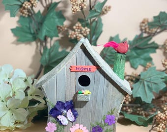 May include: A gray wooden birdhouse with a red bird on top and the word "HOME" on the front. The birdhouse is decorated with pressed flowers and a butterfly.