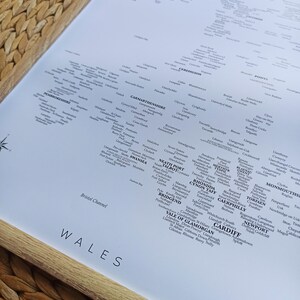 May include: A map of Wales showing the names of towns and cities in each county. The map is printed on white paper and framed in a light wood frame.