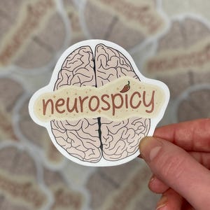 May include: A white sticker with a brain illustration and the word "neurospicy" in brown lettering. A small red chilli pepper graphic is above the word "spicy". Other stickers with brain illustrations are in the background.
