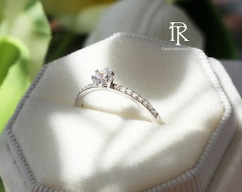 May include: A silver engagement ring with a large, clear center stone and smaller stones set along the band. The ring is displayed in a white velvet ring box. The text "Timeless Romance" is visible.