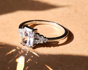 May include: A silver ring with a large, clear, rectangular-shaped gemstone surrounded by smaller, clear gemstones.