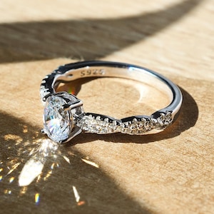 Può includere: Un anello di fidanzamento in argento con un grande diamante di forma ovale e una fascia intrecciata decorata con diamanti più piccoli.