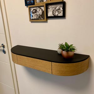 Könnte beinhalten: Ein geschwungenes Wandregal aus Holz mit einer schwarzen Oberseite und einer Schublade. Das Regal ist an einer weißen Wand montiert und hat zwei kleine Topfpflanzen auf der Oberseite.