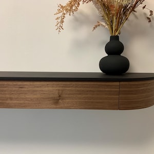 Könnte beinhalten: Ein modernes schwebendes Regal mit dunkler Oberseite und geschwungener Holzmaserungsfront. Eine schwarze Vase mit doppelter Birne enthält Trockenblumen. Das Regal ist an einer weißen Wand befestigt und schafft eine minimalistische Ästhetik.