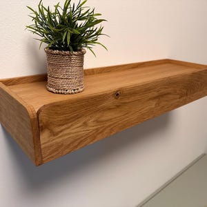Peut inclure: Étagère flottante en bois avec un grain naturel. Le bord de l'étagère est surélevé et les coins sont arrondis. Une petite plante en pot avec des feuilles vertes est posée sur l'étagère. L'étagère est fixée à un mur blanc.