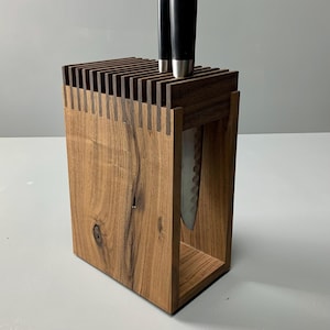 Peut inclure: Un bloc de couteaux en bois avec un fini grain de bois naturel. Le bloc a un dessus fendu pour le rangement des couteaux et une conception rectangulaire. Deux couteaux avec des manches noirs sont insérés dans le haut. Un seul couteau est visible à l'intérieur du bloc.