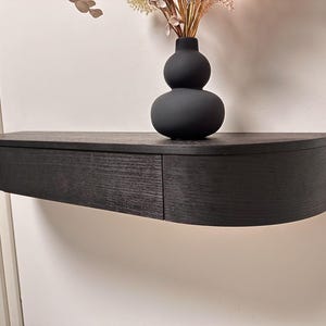Op de afbeelding: Zwarte, aan de muur gemonteerde zwevende plank met een gebogen voorkant en een lade. Een zwarte, dubbele bolvaas bevat gedroogde bloemen. De plank is gemaakt van donker hout en is gemonteerd.