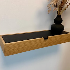 Può includere: Uno scaffale in legno con un cassetto nero e un vaso nero con fiori secchi. Lo scaffale è montato su una parete bianca.
