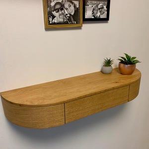 Può includere: Un ripiano a muro in legno chiaro con un fronte curvo e un unico cassetto. Il ripiano è montato su una parete bianca e ha due piccole piante in vaso sulla parte superiore.
