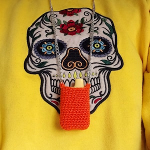 May include: A silver chain necklace with an orange crocheted pouch attached. The pouch is hanging in front of a yellow sweatshirt with a black and white embroidered sugar skull design.