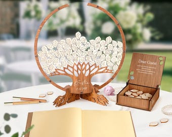 May include: A wooden guest book alternative with a tree design. The tree has a circular frame and holds small heart-shaped leaves for guests to sign. Includes a wooden box with heart-shaped tags, a guest book, and pens.