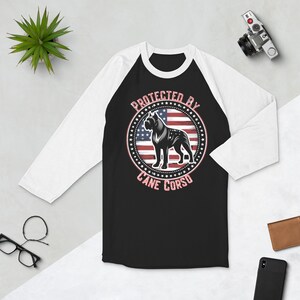 Puede incluir: Una camiseta de béisbol raglán en blanco y negro con un gráfico de un perro Cane Corso y el texto "PROTECTED BY CANE CORSO". El diseño incluye una bandera estadounidense. La camiseta tiene mangas blancas y cuerpo negro.