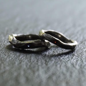 May include: Two textured silver rings with a hammered finish. One ring has a small blue gem, the other a red gem. The rings are set against a dark grey background, creating a contrast that highlights their unique design.