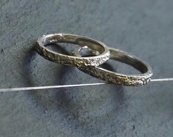 May include: Two silver rings with textured bands are shown against a grey background. The rings are close together, with one slightly overlapping the other. The bands have a rough, organic texture, suggesting a handcrafted design. These are likely wedding or engagement rings.