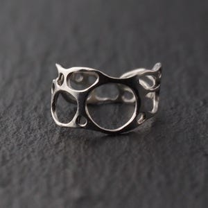 Peut inclure: Une bague en argent avec un design ajouré unique. La bague présente une série de découpes circulaires et ovales interconnectées, créant une esthétique moderne et artistique. La bague est présentée sur une surface sombre et texturée.