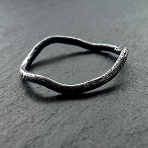 Puede incluir: Un anillo plateado con un diseño ondulado y orgánico. El anillo tiene una superficie texturizada y un acabado oscuro oxidado. La forma del anillo es irregular, lo que se suma a su apariencia única y artesanal. Es una pieza de joyería.