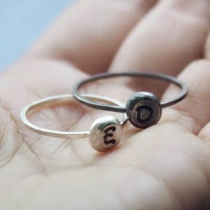 May include: Two silver rings with a round, textured band. The ring on the left has the letter "E" engraved on the band. The ring on the right has the letter "D" engraved on the band.