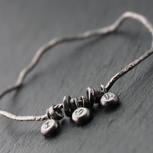 Może przedstawiać: Srebrna bransoletka typu bangle z młotkowaną fakturą. Bransoletka posiada trzy małe, okrągłe zawieszki z literami "I" i "U" oraz symbolem serca. Bransoletka leży na ciemnoszarej powierzchni.