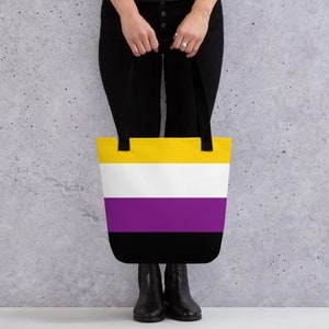 Può includere: Una borsa tote con un designo a strisce gialle, bianche, viola e nere. La borsa ha manici neri ed è tenuta da una persona.