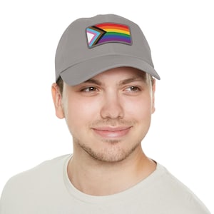 May include: A gray baseball cap featuring a colorful patch with a rainbow and other LGBTQ+ pride flag symbols. The cap is made of a soft material and has a curved brim. The cap is a stylish accessory.