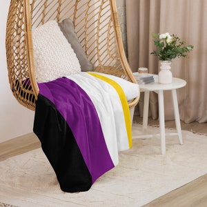 May include: A purple, white, black, and yellow striped throw blanket draped over a wicker chair. The blanket is a representation of the non-binary pride flag.