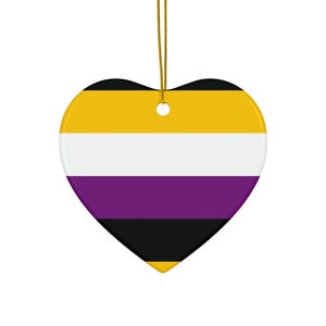 Puede incluir: Un adorno de cerámica en forma de corazón con el diseño de la bandera del orgullo no binario, que presenta franjas horizontales de color amarillo, blanco, morado y negro. El adorno incluye un pequeño orificio superior y un cordón dorado.