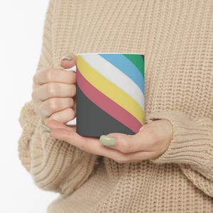 Könnte beinhalten: Ein Keramikbecher mit einem diagonalen Streifenmuster in den Farben Dunkelgrau, gedämpftes Rot, Gelb, Weiß, Hellblau und Grün. Der Becher hat einen glatten, zylindrischen Körper und einen einfachen Henkel, was ein klares Design ergibt.