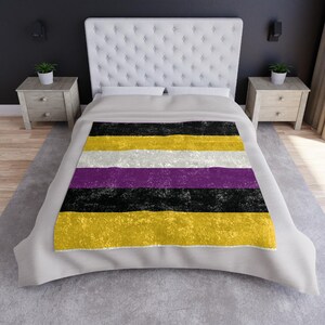 May include: A non-binary pride flag blanket with horizontal stripes of yellow, white, purple, black, and yellow. The blanket is displayed on a bed with a white tufted headboard and two wooden nightstands.