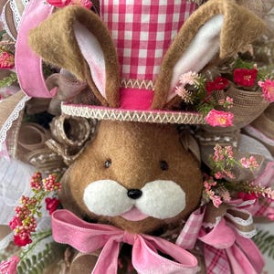 May include: A decorative Easter wreath featuring a brown bunny face with a pink bow and a gingham top hat. The wreath is adorned with pink and white ribbons, faux flowers, and burlap accents, creating a festive spring theme.