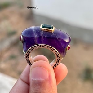 Op de afbeelding: Een sierlijke ring met een gepolijste, dieppaarse steen. De ring heeft een zilveren band met een zich herhalend cirkelontwerp en is geaccentueerd met een kleine groene steen en twee rode stenen. Het woord "Ronak" staat in de linkerbovenhoek.