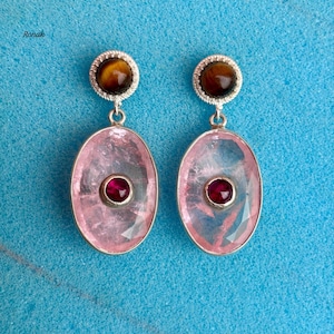 Op de afbeelding: Een paar zilveren oorbellen met een tijgeroogsteen aan de bovenkant en een grote, ovale, roze edelsteen met een kleinere, ronde, rode edelsteen in het midden. De oorbellen hangen aan een zilveren haak tegen een blauwe achtergrond.