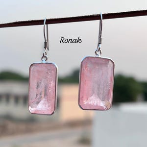 May include: A pair of rectangular pink gemstone earrings with silver-colored metal frames and hooks. The earrings are hanging from a dark brown rod. The word "Ronak" is printed in black on the right side of the image.