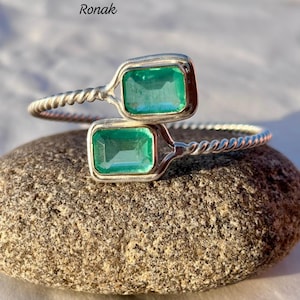 Op de afbeelding: Een zilveren armband met twee smaragdgroene edelstenen. De armband heeft een gedraaid ontwerp.