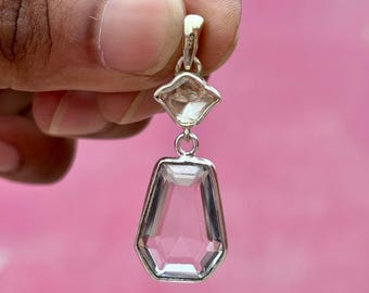 Può includere: Un pendente trasparente e sfaccettato con montatura color argento. Il pendente ha un design geometrico con una pietra più piccola a forma di diamante sopra una pietra rettangolare più grande. La parola "Ronak" è nell'angolo in alto a sinistra.