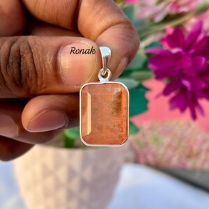 May include: A rectangular pendant with a silver bail and a reddish-orange gemstone. The gemstone has a textured appearance and is set in a silver frame. The word "Ronak" is visible on the pendant.