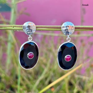 Peut inclure: Paire de boucles d'oreilles en argent avec une partie supérieure martelée. Chaque boucle d'oreille présente une grande pierre noire ovale avec une petite pierre rouge ronde au centre. Arrière-plan flou.
