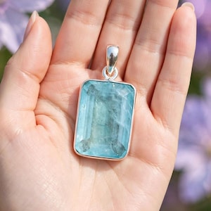 Puede incluir: Un colgante de piedra preciosa azul claro, rectangular y facetado, engastado en un marco de plata pulida. La piedra presenta un aspecto claro y translúcido con inclusiones internas visibles y una superficie lisa y reflectante. Tiene una anilla de plata.
